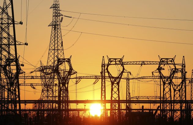 Torres e fios elétricos em silhueta contra um céu laranja e dourado, com o sol brilhando no horizonte.
