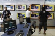 Três homens em uma loja de TVs: um segurança armado, um funcionário ajustando telas e um cliente observando.