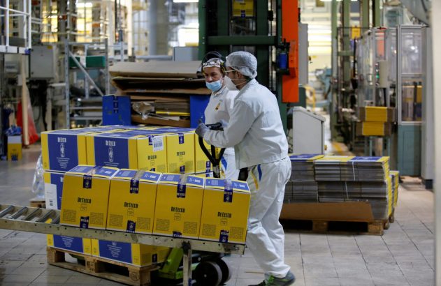 Um trabalhador empurra um carrinho de paletes com caixas De Cecco em uma fábrica, outro trabalhador observa.