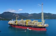 Navio FPSO com complexa estrutura amarela e vermelha, flutua em águas azuis, com montanhas verdes ao fundo. Barcos próximos.