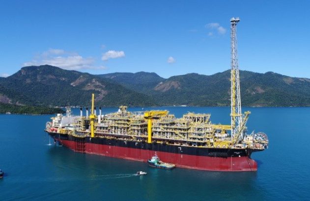 Navio FPSO com complexa estrutura amarela e vermelha, flutua em águas azuis, com montanhas verdes ao fundo. Barcos próximos.