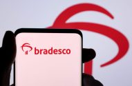 Mão segura smartphone exibindo logo Bradesco em vermelho. O mesmo logo aparece desfocado no fundo.