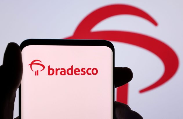Mão segura smartphone exibindo logo Bradesco em vermelho. O mesmo logo aparece desfocado no fundo.