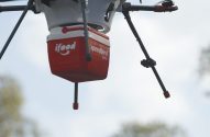 Drone branco carrega caixa vermelha iFood/Speedbird voando sob céu claro.