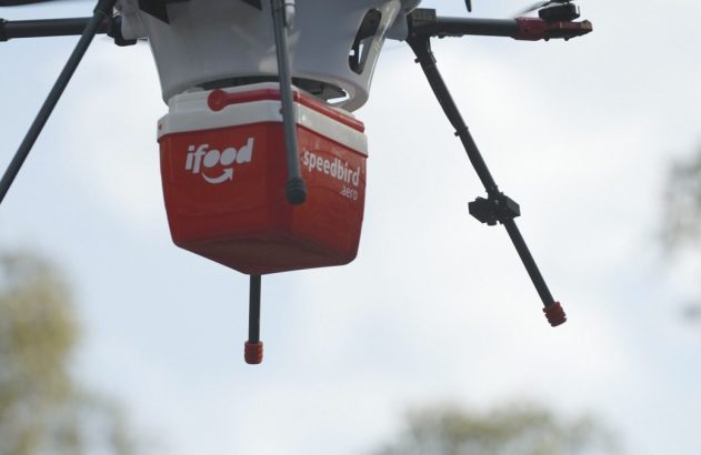 Drone branco carrega caixa vermelha iFood/Speedbird voando sob céu claro.