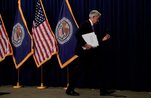 Presidente do Fed caminhando com papéis, bandeiras dos EUA e Fed ao fundo.