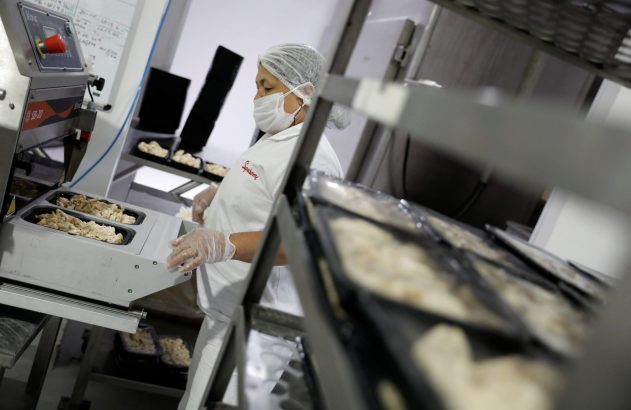 Funcionária em cozinha industrial, usando equipamento de proteção, embala refeições prontas em bandejas com máquina.