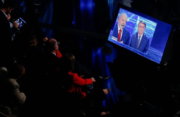 Pessoas assistem a debate presidencial entre Lula e Bolsonaro em tela grande; um com tablet.