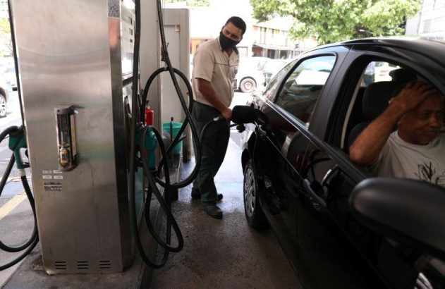Frentista mascarado abastece carro preto; motorista no veículo com expressão preocupada.
