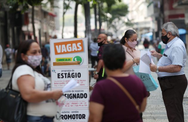 Pessoas com máscaras em rua movimentada. Um homem entrega documentos a uma mulher ao lado de placa 'VAGAS' de emprego.