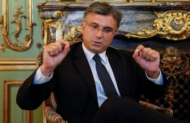 Homem de terno gesticulando com as mãos, sentado em cadeira ornamentada num ambiente clássico.