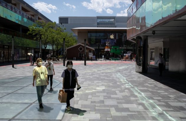 Pessoas com máscaras andam em área de compras moderna ao ar livre, com lojas Apple e Dior, sob céu ensolarado.