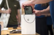 Mãos colocam um iPhone verde com câmeras na sacola branca da Apple com o logo, sobre mesa de madeira.