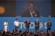 Sergio Massa discursa em palco azul com outros políticos e projeção em tela, diante de uma multidão em comício.