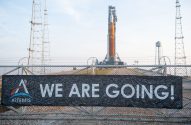 Banner "WE ARE GOING!" em cerca, com foguete Artemis SLS em plataforma de lançamento ao fundo.