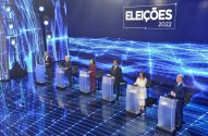 Debate das Eleições 2022. Seis candidatos em pódios num palco com iluminação azul e gráficos digitais.