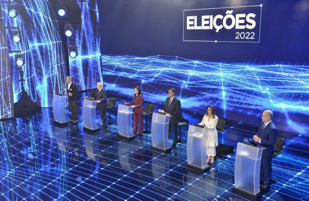 Debate das Eleições 2022. Seis candidatos em pódios num palco com iluminação azul e gráficos digitais.