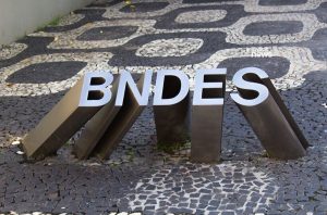 Letreiro BNDES prateado sobre calçada de pedras portuguesas preta e branca com padrão sinuoso.