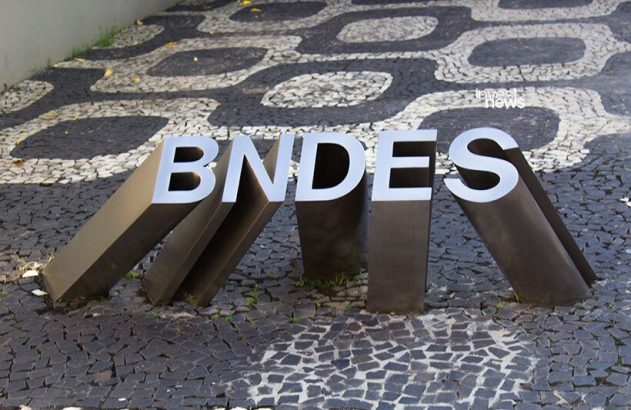 Letreiro BNDES prateado sobre calçada de pedras portuguesas preta e branca com padrão sinuoso.