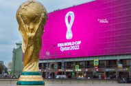 Troféu dourado da Copa do Mundo FIFA com tela grande rosa exibindo 'FIFA World Cup Qatar 2022' ao fundo.