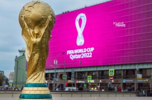 Troféu dourado da Copa do Mundo FIFA com tela grande rosa exibindo 'FIFA World Cup Qatar 2022' ao fundo.