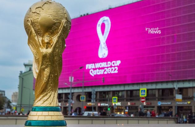 Troféu dourado da Copa do Mundo FIFA com tela grande rosa exibindo 'FIFA World Cup Qatar 2022' ao fundo.