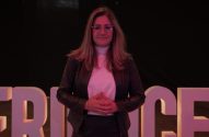 Mulher de óculos e mãos unidas sorrindo em frente a letras iluminadas.