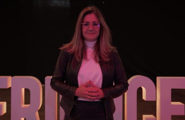 Mulher de óculos e mãos unidas sorrindo em frente a letras iluminadas.