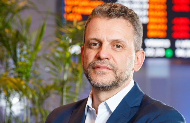 Homem com barba e terno, olhando para a câmera, em frente a uma tela de dados numéricos e plantas.