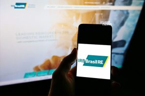 Mão segura smartphone exibindo o logo IRB Brasil RE. Monitor com site da empresa no segundo plano.