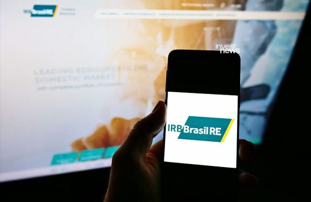 Mão segura smartphone exibindo o logo IRB Brasil RE. Monitor com site da empresa no segundo plano.