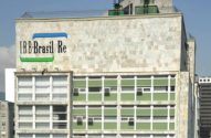 Prédio cinza com logo 'IRB Brasil Re' (barras azul e verde) no topo. Janelas com molduras brancas e painéis verdes.