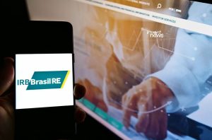 Mão segura celular com logo IRB Brasil RE. Monitor no fundo mostra site financeiro 'invest news' e gráficos.