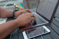 Mãos digitando em laptop com planilha financeira. Celular ao lado exibe gráfico de bolsa de valores.