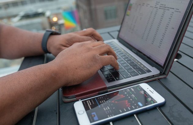 Mãos digitando em laptop com planilha financeira. Celular ao lado exibe gráfico de bolsa de valores.