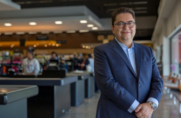Homem de terno azul e óculos posa sorrindo em um supermercado, com caixas registradoras desfocadas ao fundo.