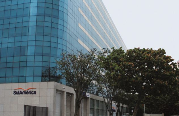 Edifício moderno de fachada azul-espelhada com o logo SulAmérica e árvores no primeiro plano.