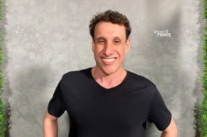 Um homem sorrindo amplamente, com cabelo cacheado e camiseta preta, em frente a uma parede cinza texturizada.