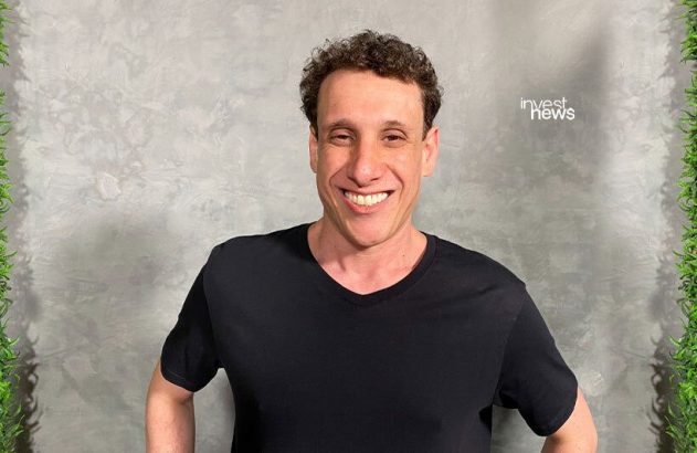 Um homem sorrindo amplamente, com cabelo cacheado e camiseta preta, em frente a uma parede cinza texturizada.