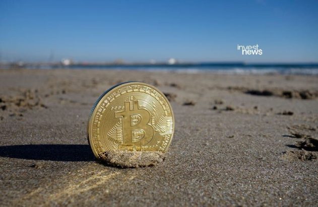 Uma moeda Bitcoin dourada fincada na areia da praia, com o mar ao fundo.
