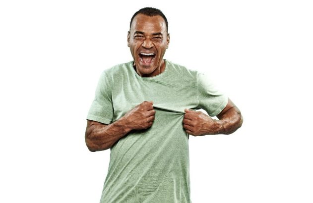 Homem de camiseta verde, rindo e puxando a própria camisa com as mãos.