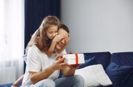 Filha surpreende pai com presente, cobrindo seus olhos enquanto ele segura a caixa, ambos sorrindo.