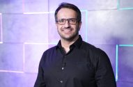 Homem sorrindo, de óculos e camisa preta. Fundo geométrico com luzes neon coloridas.