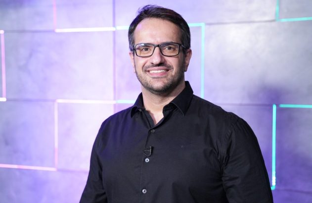 Homem sorrindo, de óculos e camisa preta. Fundo geométrico com luzes neon coloridas.