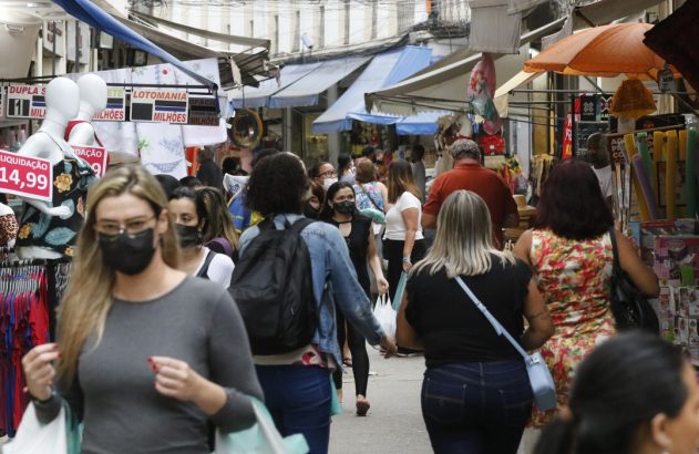 Rua comercial movimentada com pedestres, muitos de máscara, e barracas de produtos diversos.