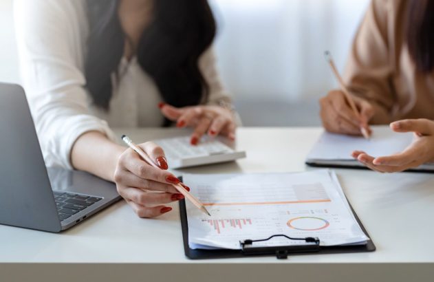 A imagem mostra um iPhone branco exibindo um gráfico de cotação de ações. O celular está em cima de uma folha de papel em que foi impressa uma tabela com vários dados. (Foto: Pexels / Olya Kobruseva)