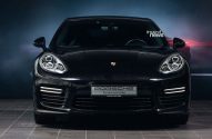 Porsche Panamera preto, vista frontal, em showroom com luzes diurnas acesas.