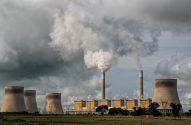 Usina de energia com grandes torres de resfriamento e chaminés, uma emitindo fumaça densa sob céu nublado.