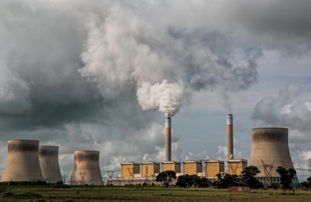Usina de energia com grandes torres de resfriamento e chaminés, uma emitindo fumaça densa sob céu nublado.
