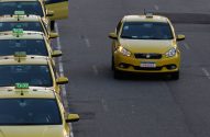 Fila de táxis amarelos estacionados à esquerda e um táxi amarelo em movimento na pista.